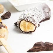 A frozen banana half on a stick, partially coated in chocolate and sprinkled with shredded coconut, rests on a white surface next to pieces of chocolate – a simple frozen bananas recipe for a delicious banana dessert.