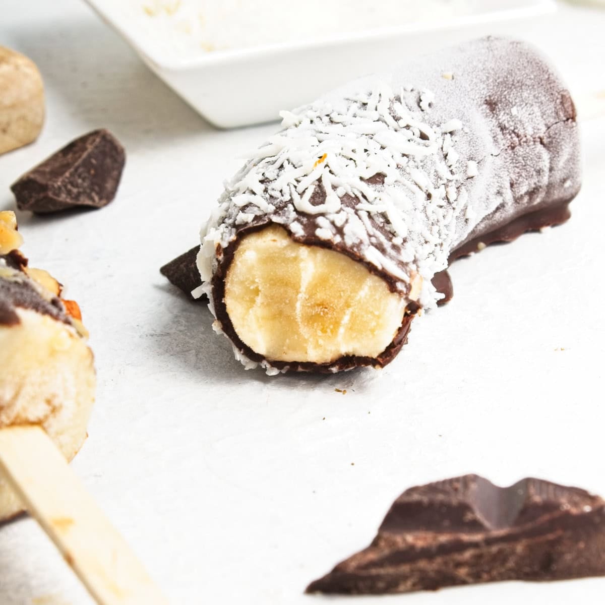 A frozen banana half on a stick, partially coated in chocolate and sprinkled with shredded coconut, rests on a white surface next to pieces of chocolate – a simple frozen bananas recipe for a delicious banana dessert.
