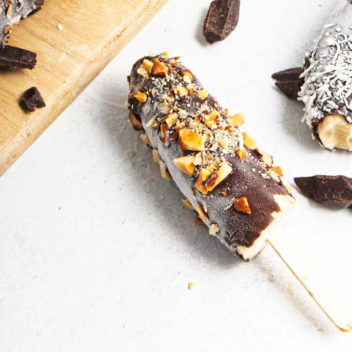 Chocolate covered banana popsicle topped with chopped nuts, placed on a white surface with chocolate chunks and another popsicle nearby—a delightful banana dessert and a classic frozen bananas recipe.