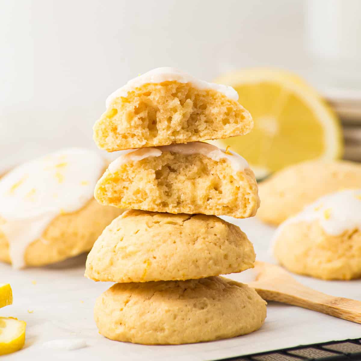 A stack of lemon cookies with glaze, two cookies broken in half to show the soft interior, with a lemon slice and more cookies in the background.