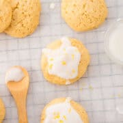 Round lemon cookies with white icing and lemon zest are arranged on a grid-patterned cloth, next to a wooden spoon with icing and a small glass bowl of icing.