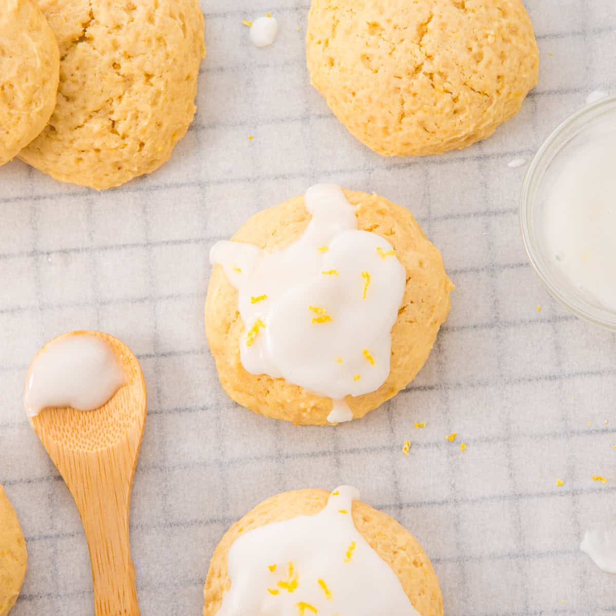 Round lemon cookies with white icing and lemon zest are arranged on a grid-patterned cloth, next to a wooden spoon with icing and a small glass bowl of icing.