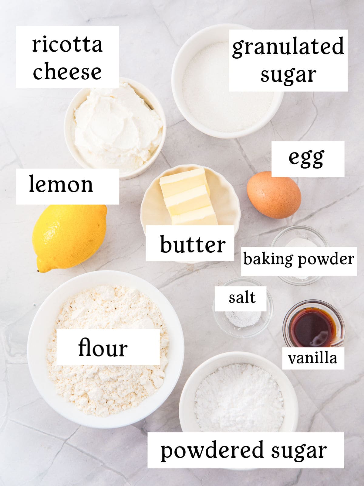 Top-down view of labeled baking ingredients: ricotta cheese, granulated sugar, egg, lemon, butter, flour, baking powder, salt, vanilla, and powdered sugar on a light surface.