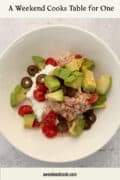 A white bowl containing tuna, diced avocado, cherry tomatoes, black olives, and cottage cheese on a light surface.