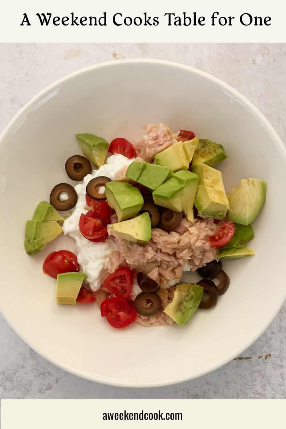 A white bowl containing tuna, diced avocado, cherry tomatoes, black olives, and cottage cheese on a light surface.