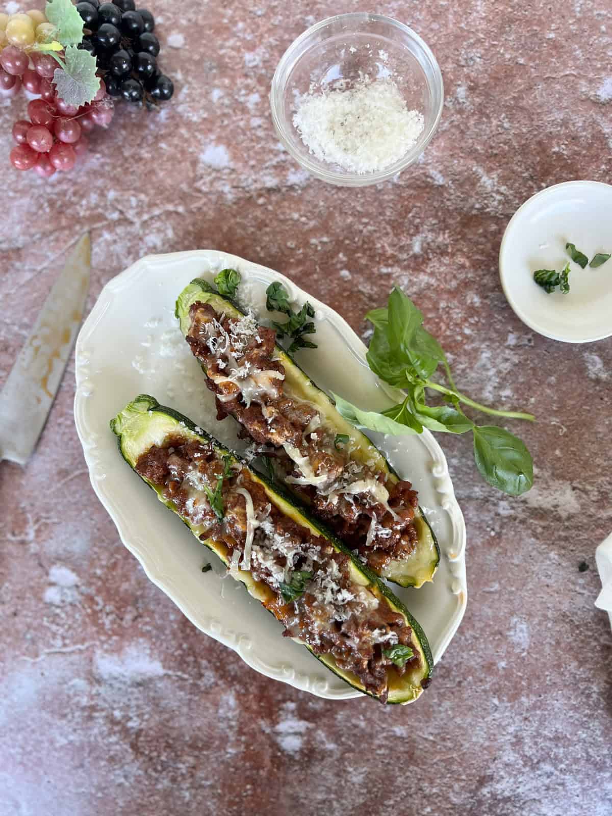 Two zucchini boats filled with a ground meat mixture, topped with grated cheese and herbs, served on a white oval plate with fresh basil. Grated cheese, grapes, and a knife are nearby.