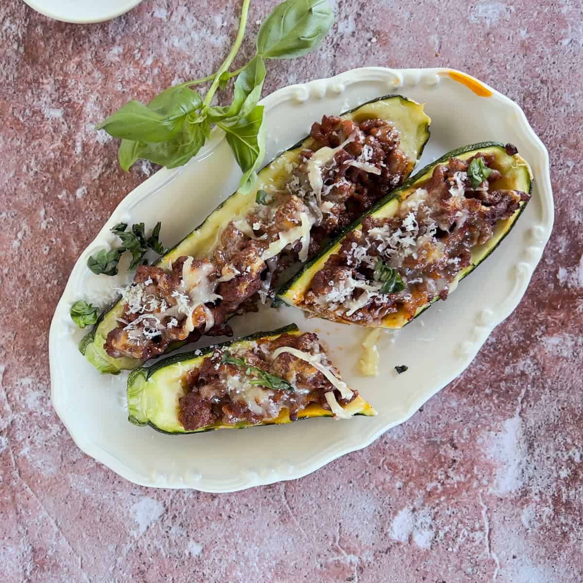 Three zucchini boats filled with ground meat, herbs, and topped with grated cheese are arranged on a white decorative platter, garnished with fresh basil.