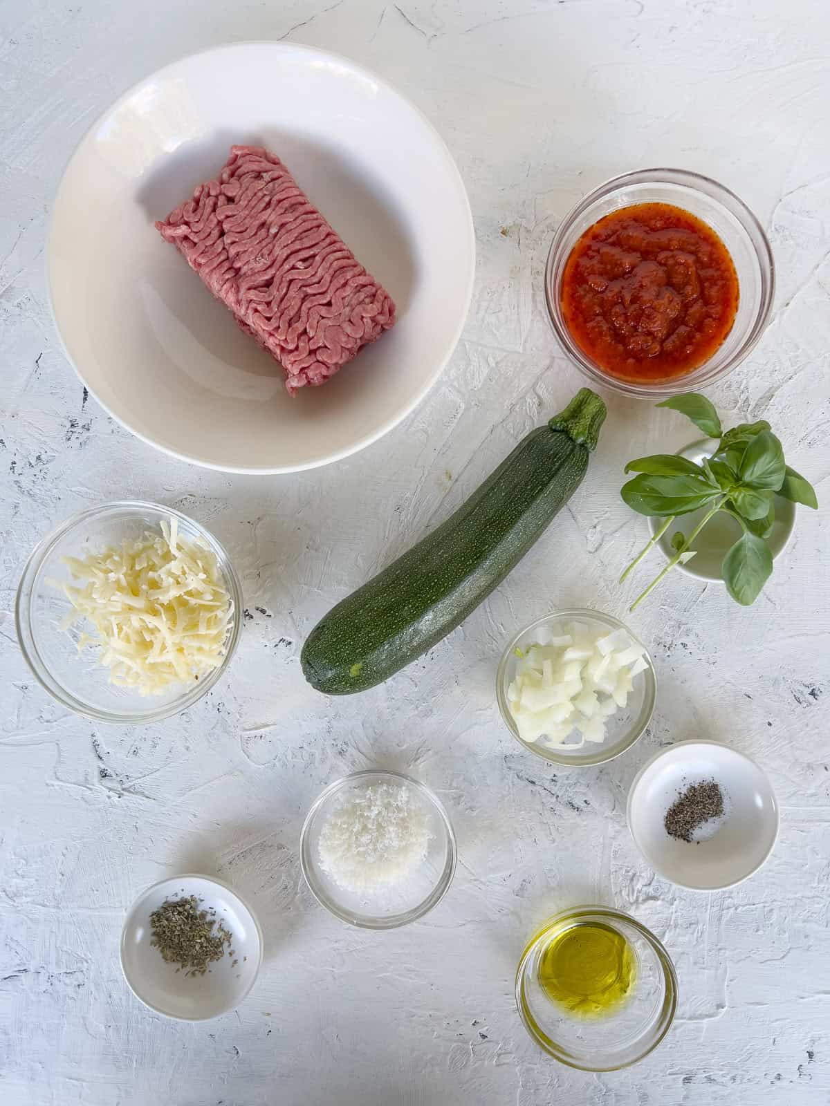 Ingredients laid out on a white surface for zucchini boats, including ground meat, tomato sauce, zucchini, basil, grated cheeses, chopped onion, olive oil, salt, pepper, and dried herbs in small bowls.
