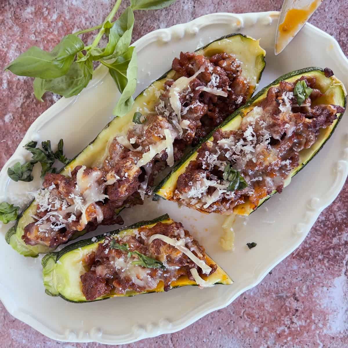 Three zucchini boats filled with a ground meat and tomato mixture, topped with shredded cheese and fresh herbs, served on a white plate.