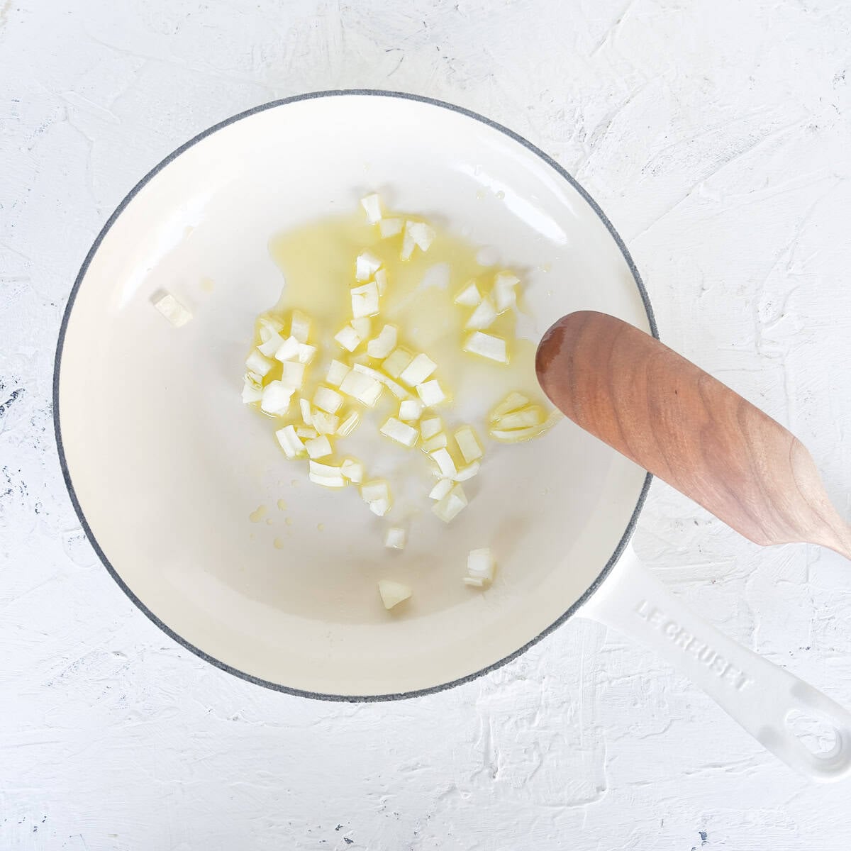 Chopped onions sautéing in oil in a white pot with a wooden spoon—perfect for starting your zucchini boats—on a white textured surface.