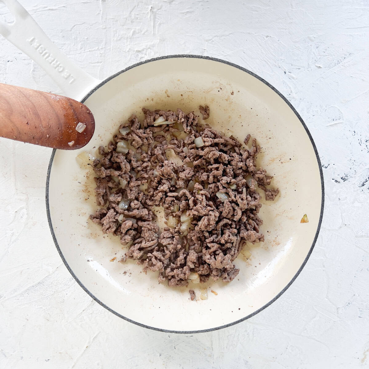 Ground beef and chopped onions cooking in a white skillet with a wooden handle, perfect for stuffing into zucchini boats, set on a textured white surface.