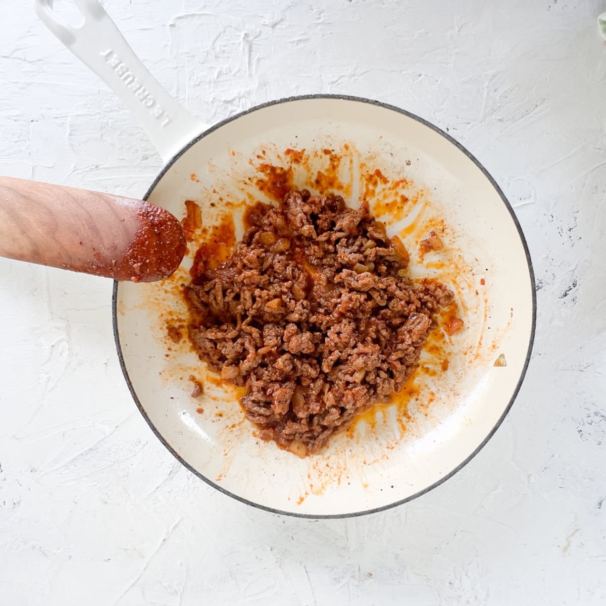 A white skillet containing cooked, seasoned ground beef for zucchini boats sits on a white surface, with a wooden spatula partially covered in sauce resting on the pan's edge.