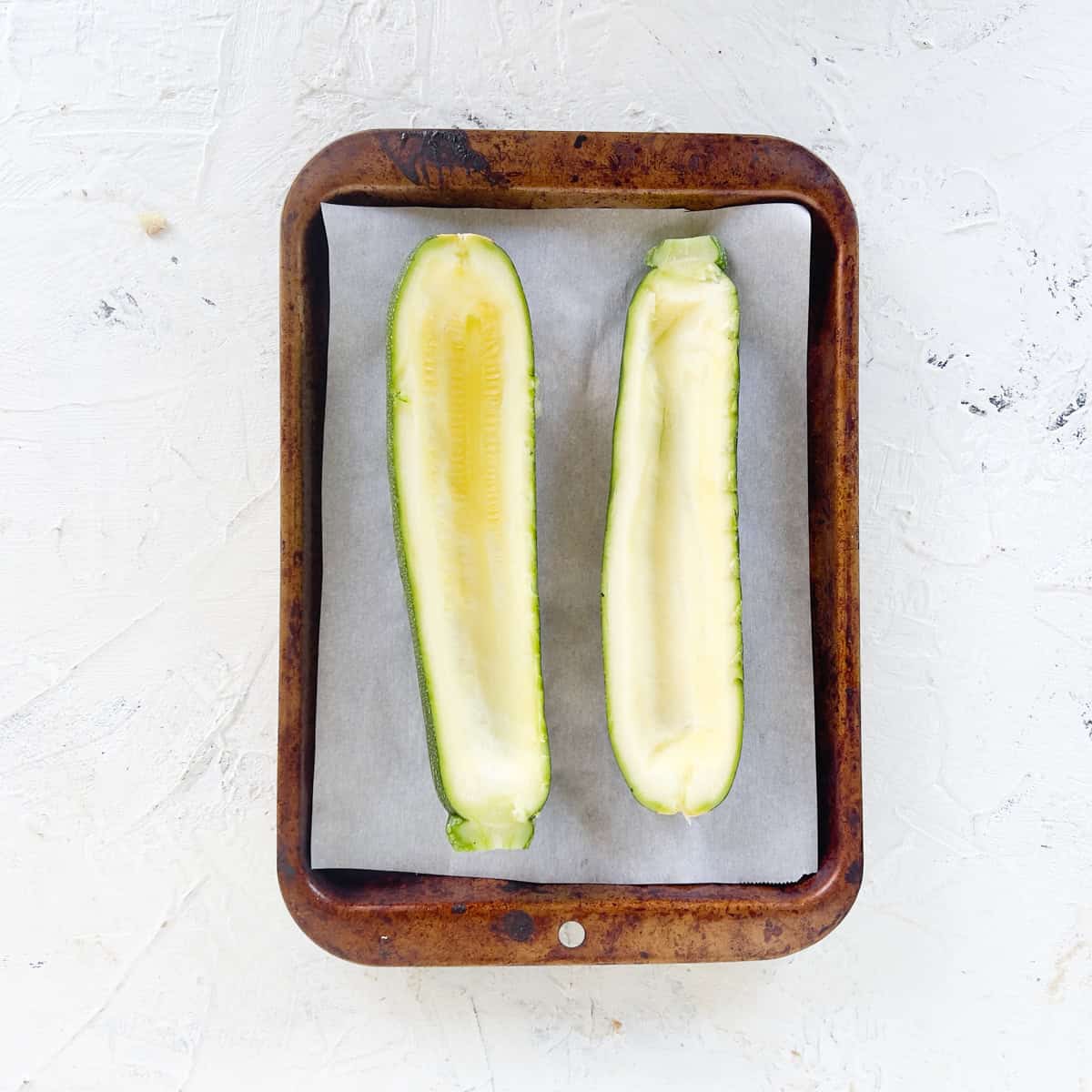 A baking tray lined with parchment paper holds two zucchini boats with the centers scooped out, placed side by side on a white textured surface.