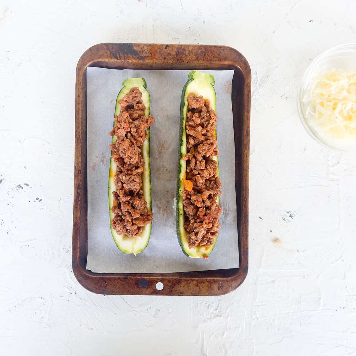 Two zucchini boats filled with cooked ground meat mixture rest on a baking tray lined with parchment paper; a bowl of shredded cheese sits close by.