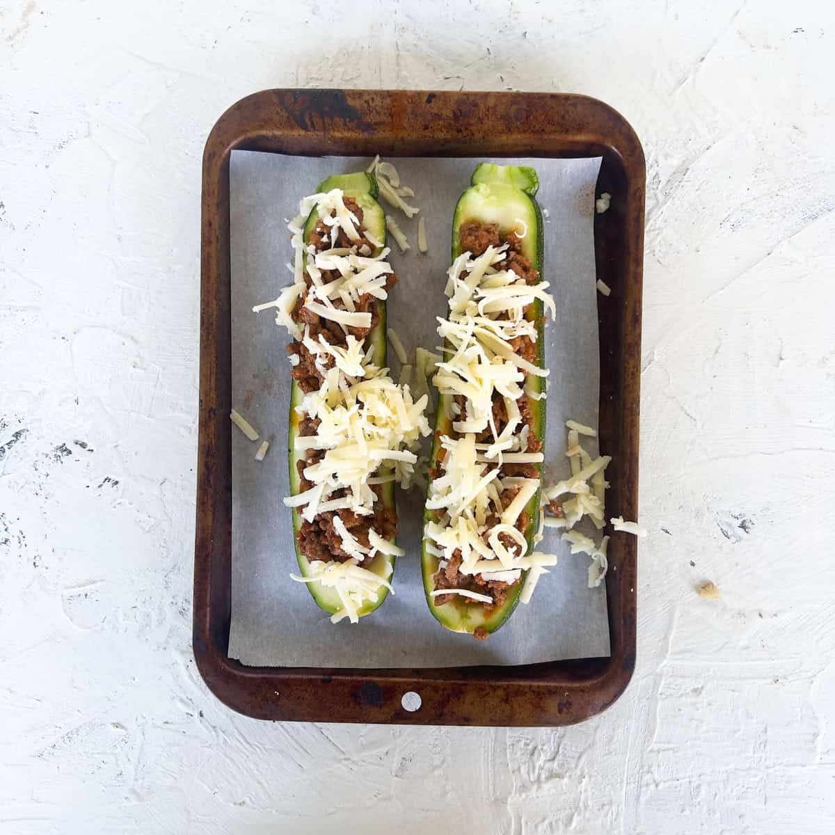 Two zucchini boats filled with a savory meat mixture and topped with shredded cheese rest on parchment paper in a baking tray.