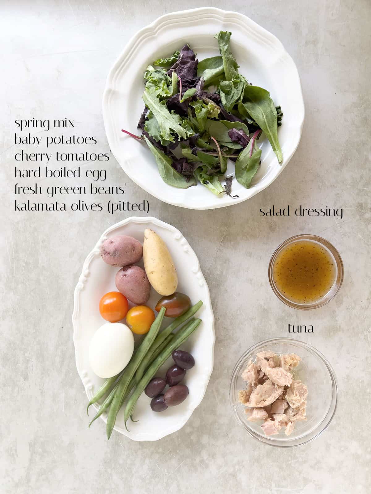 Three plates with spring mix, baby potatoes, cherry tomatoes, hard boiled egg, green beans, kalamata olives, salad dressing, and tuna, arranged on a light surface with labels.
