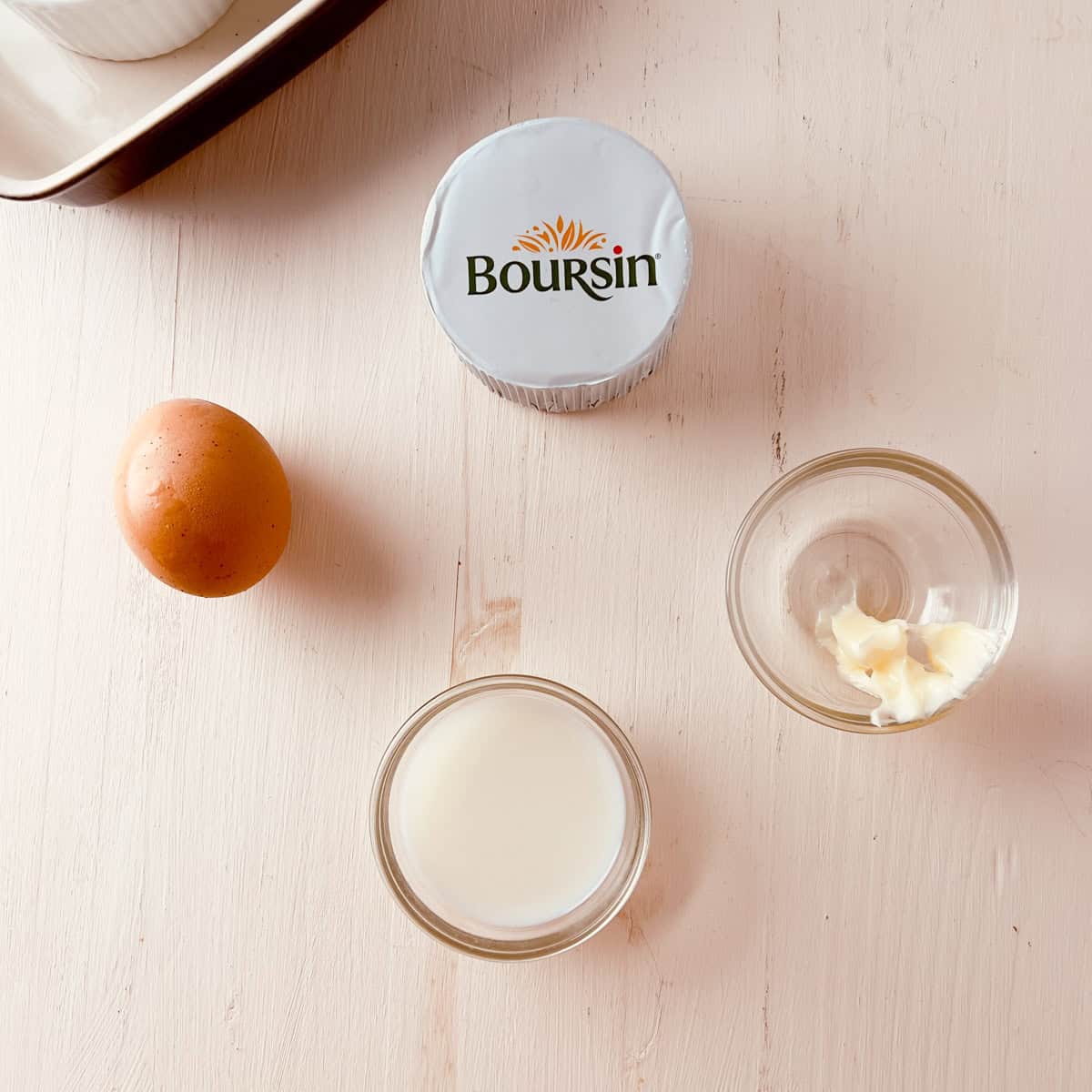 A brown egg, a small glass of milk, a small dish of butter, and a sealed container of Boursin cheese on a light wooden surface.