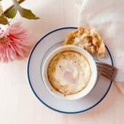 A baked egg dish in a white ramekin sits on a white plate with a blue rim, alongside a piece of bread, a fork, a pink flower, and a white cloth on a light wooden surface.