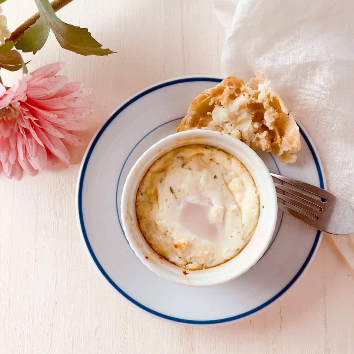 A baked egg dish in a white ramekin sits on a white plate with a blue rim, alongside a piece of bread, a fork, a pink flower, and a white cloth on a light wooden surface.