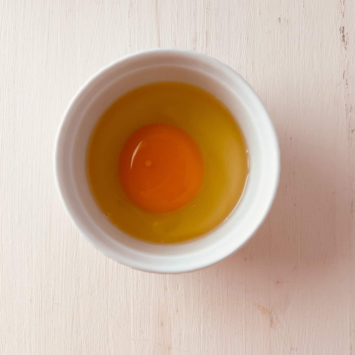 A raw egg with an intact yolk in a white bowl, placed on a light wooden surface.