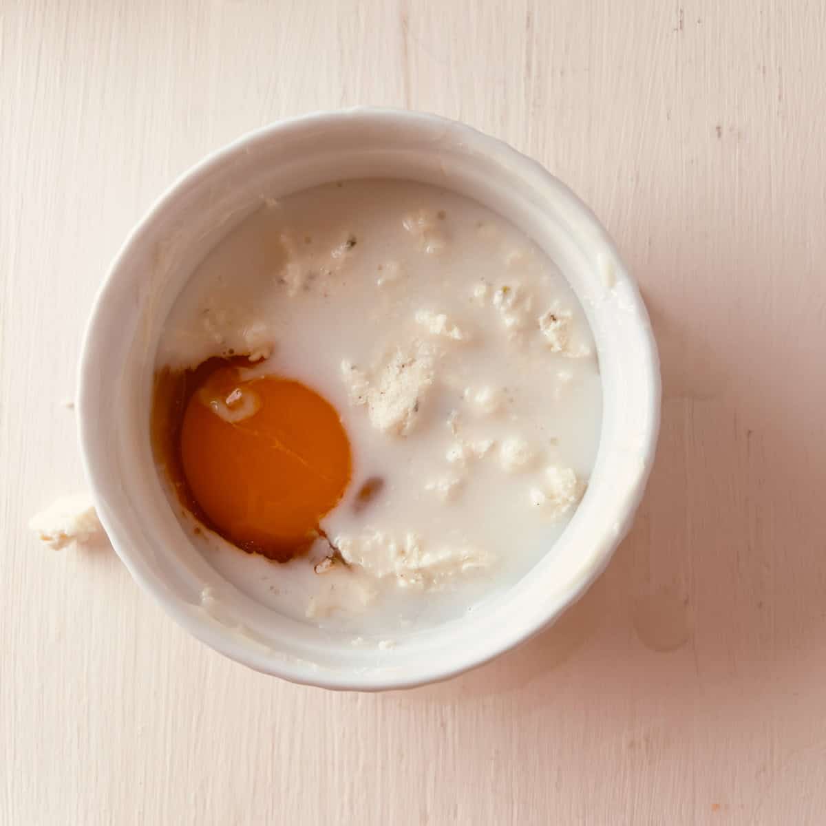 A white bowl containing an egg yolk, egg white, and milk mixture, with some lumps, sits on a light-colored wooden surface.
