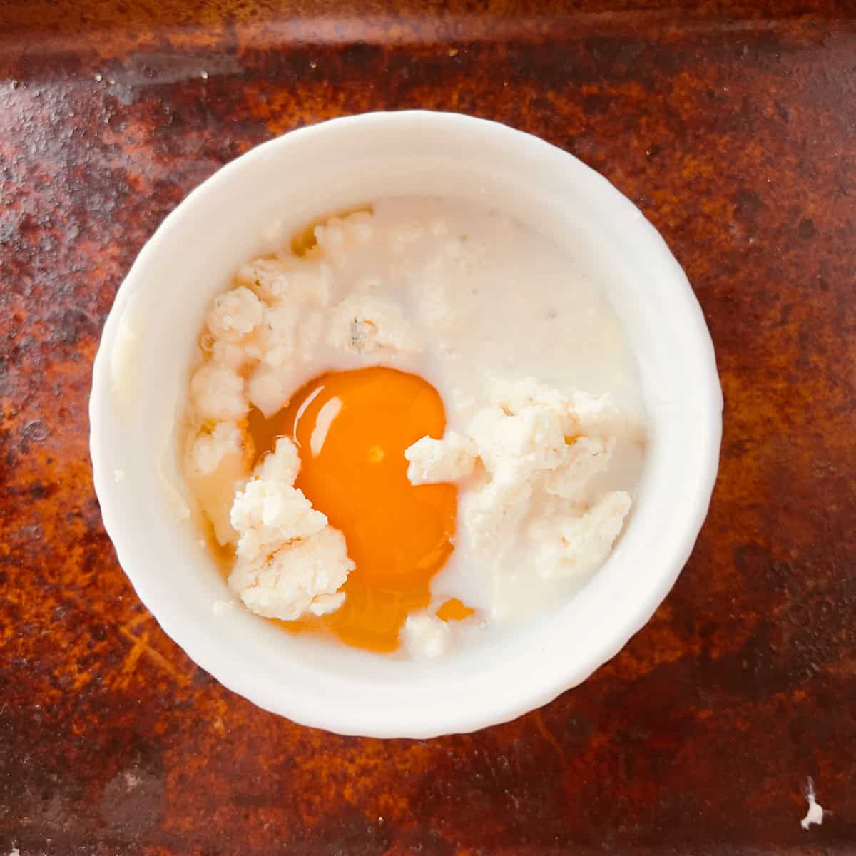 A raw egg and crumbled cheese in a small white ramekin on a brown textured surface.