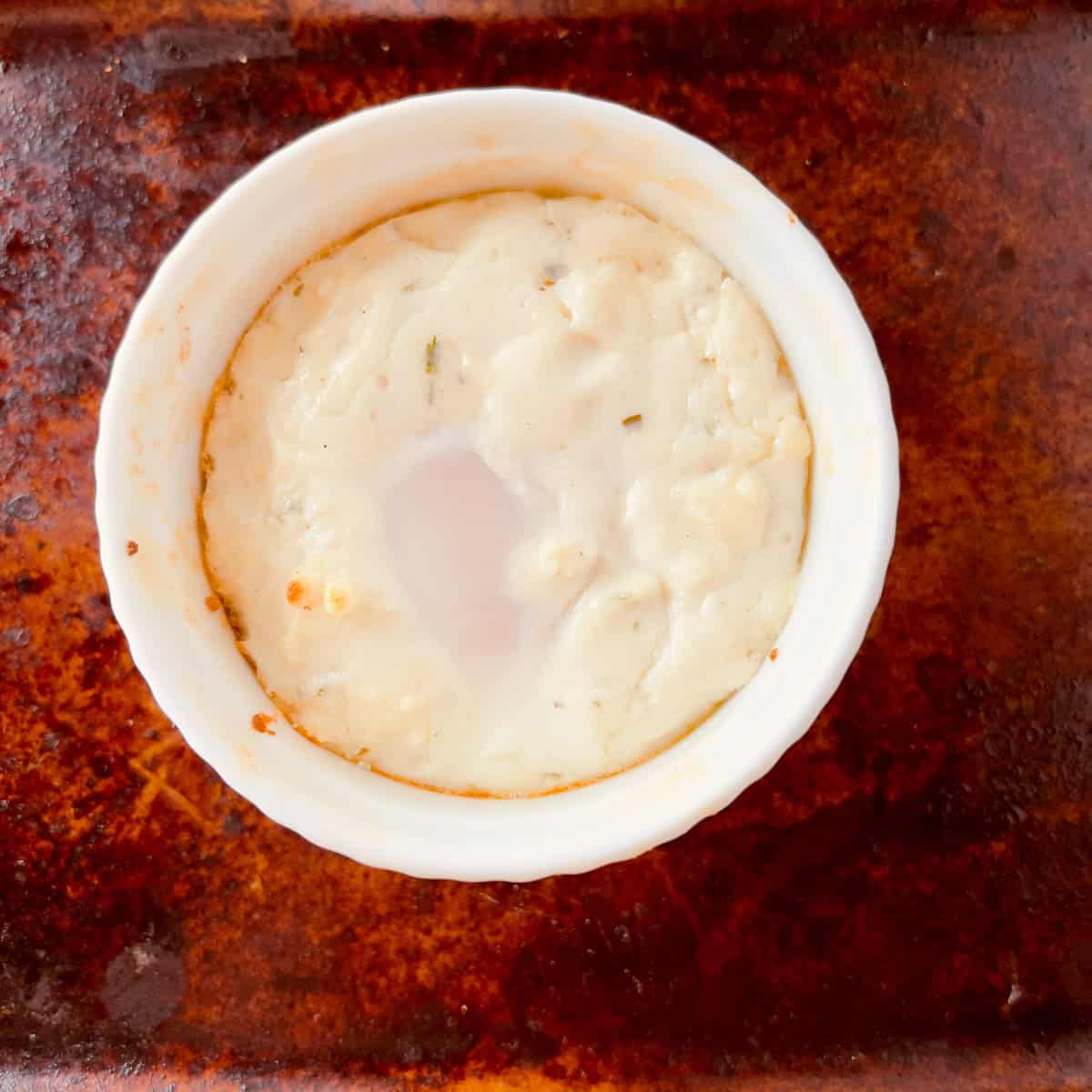 A baked egg dish in a white ramekin sits on a brown, textured surface. The egg is set with a slightly soft center and a lightly browned edge.