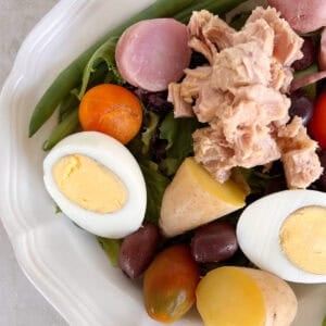 A plate with green beans, hard-boiled eggs, cherry tomatoes, olives, boiled potatoes, mixed greens, and a serving of tuna.