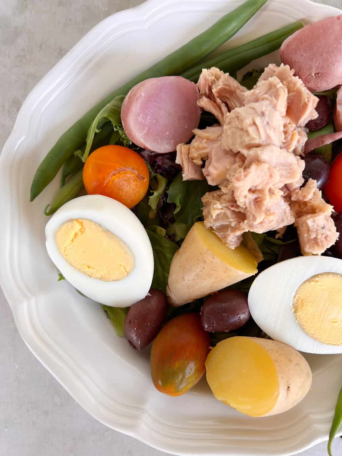 A plate with green beans, hard-boiled eggs, cherry tomatoes, olives, boiled potatoes, mixed greens, and a serving of tuna.