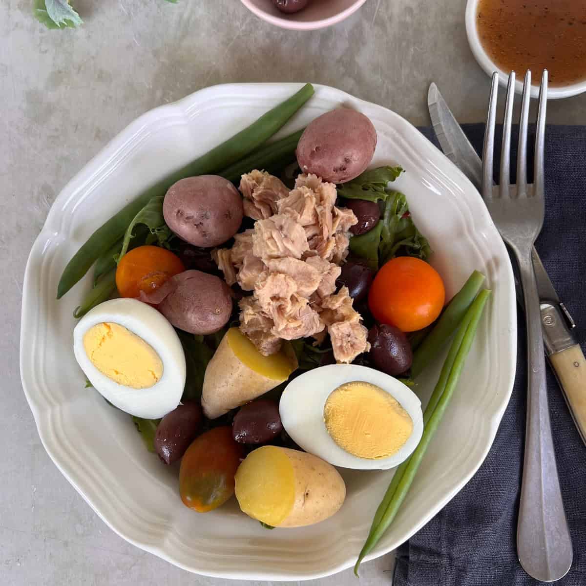 A plate of salad with hard-boiled eggs, baby potatoes, cherry tomatoes, green beans, tuna, and olives on mixed greens. Fork, knife, and dressing cup are beside the plate.