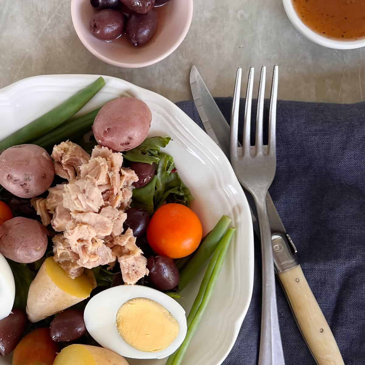 A nicoise salad with tuna, boiled eggs, cherry tomatoes, green beans, red potatoes, and olives, next to a fork, knife, small bowl of olives, and a cup of dressing.