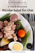 A plate of Nicoise salad featuring tuna, hard-boiled egg, cherry tomato, green beans, potatoes, and olives, with a fork and knife placed beside the dish.