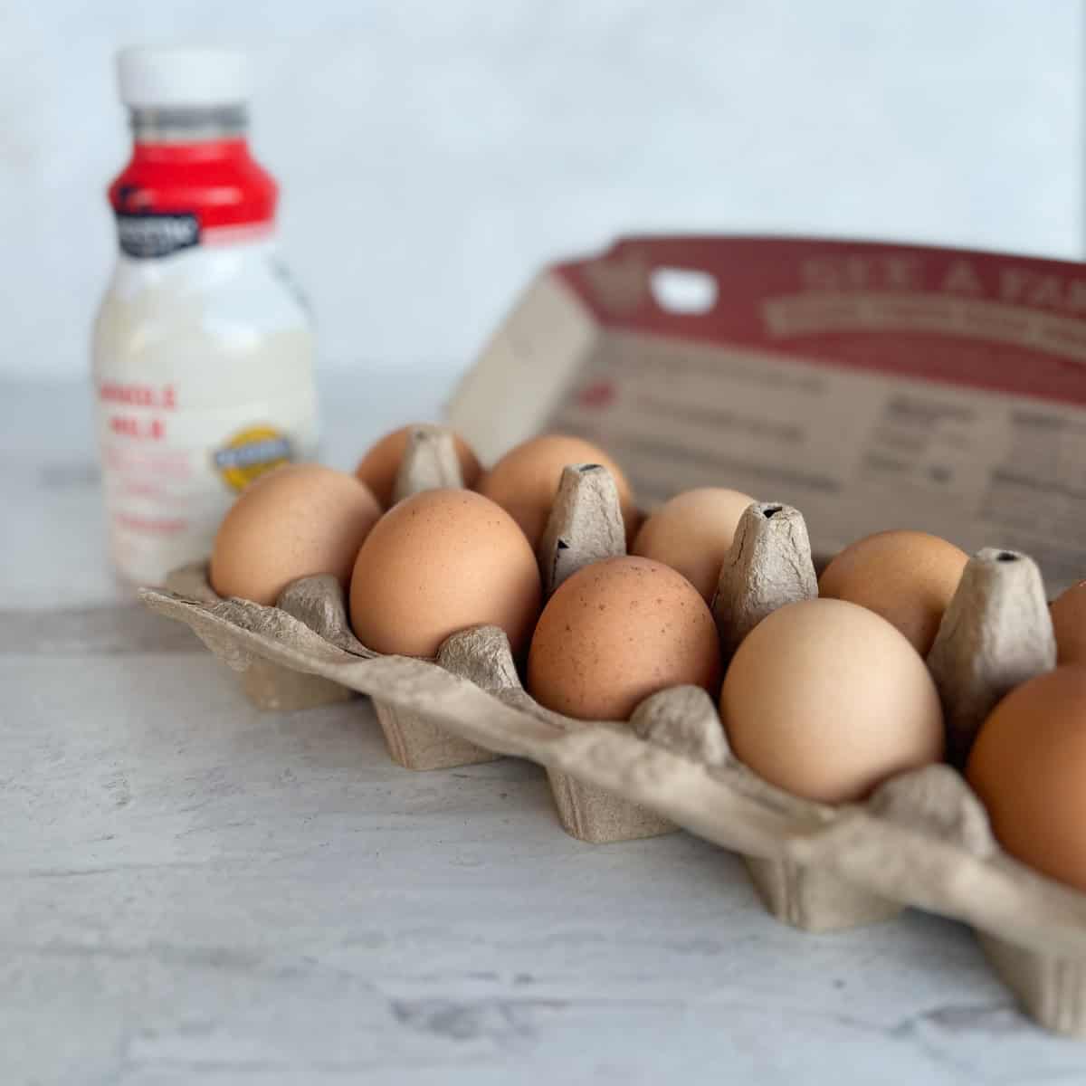 A carton of brown eggs sits on a light surface with a bottle of buttermilk in the background.