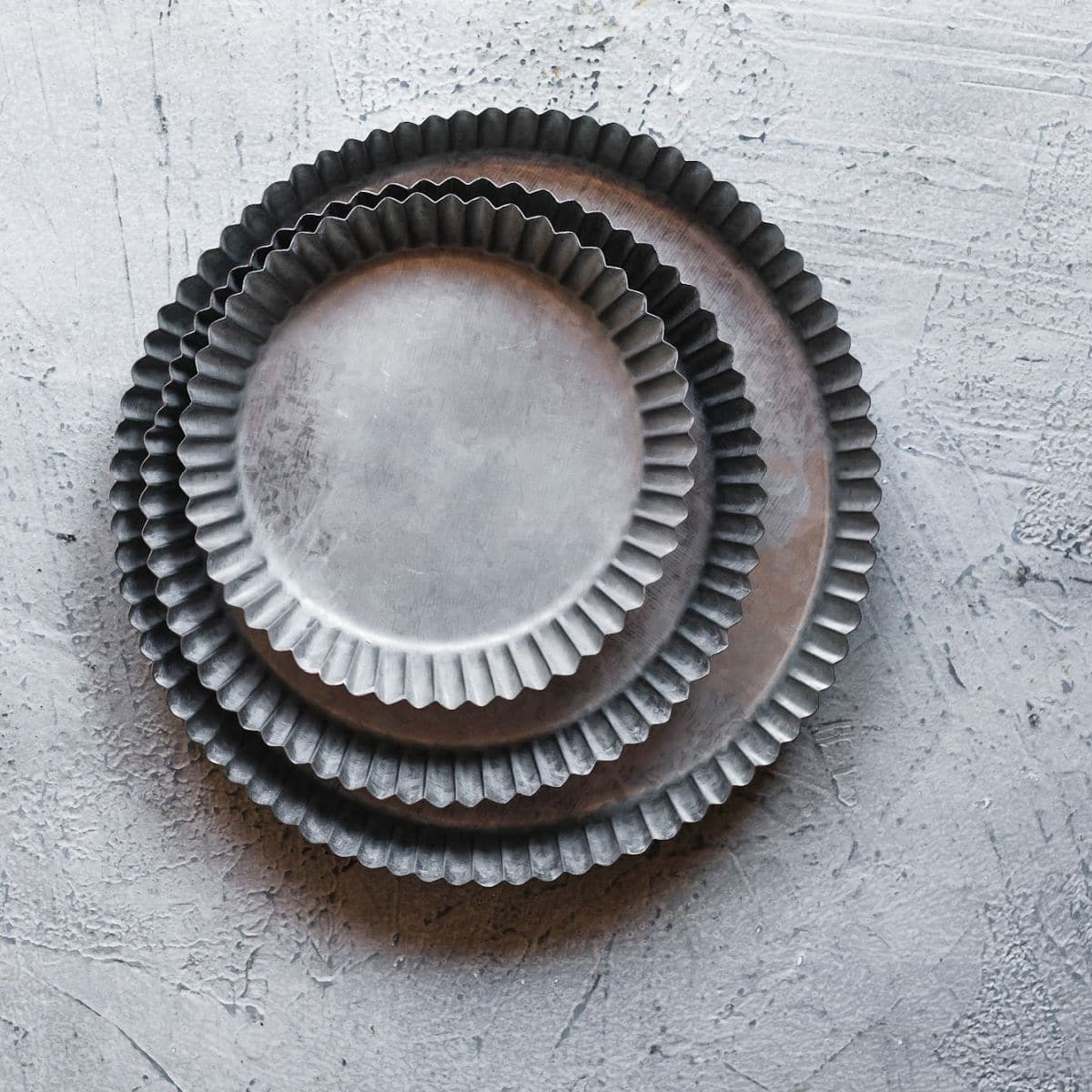 Three round, fluted metal tart pans nested together on a textured grey surface—perfect for baking a small batch quiche or making the best pan choice when preparing quiche for one.