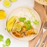 Plate with rice, grilled chicken topped with a lime slice and cilantro, avocado slices, a fork, a knife, and a brown napkin nearby.