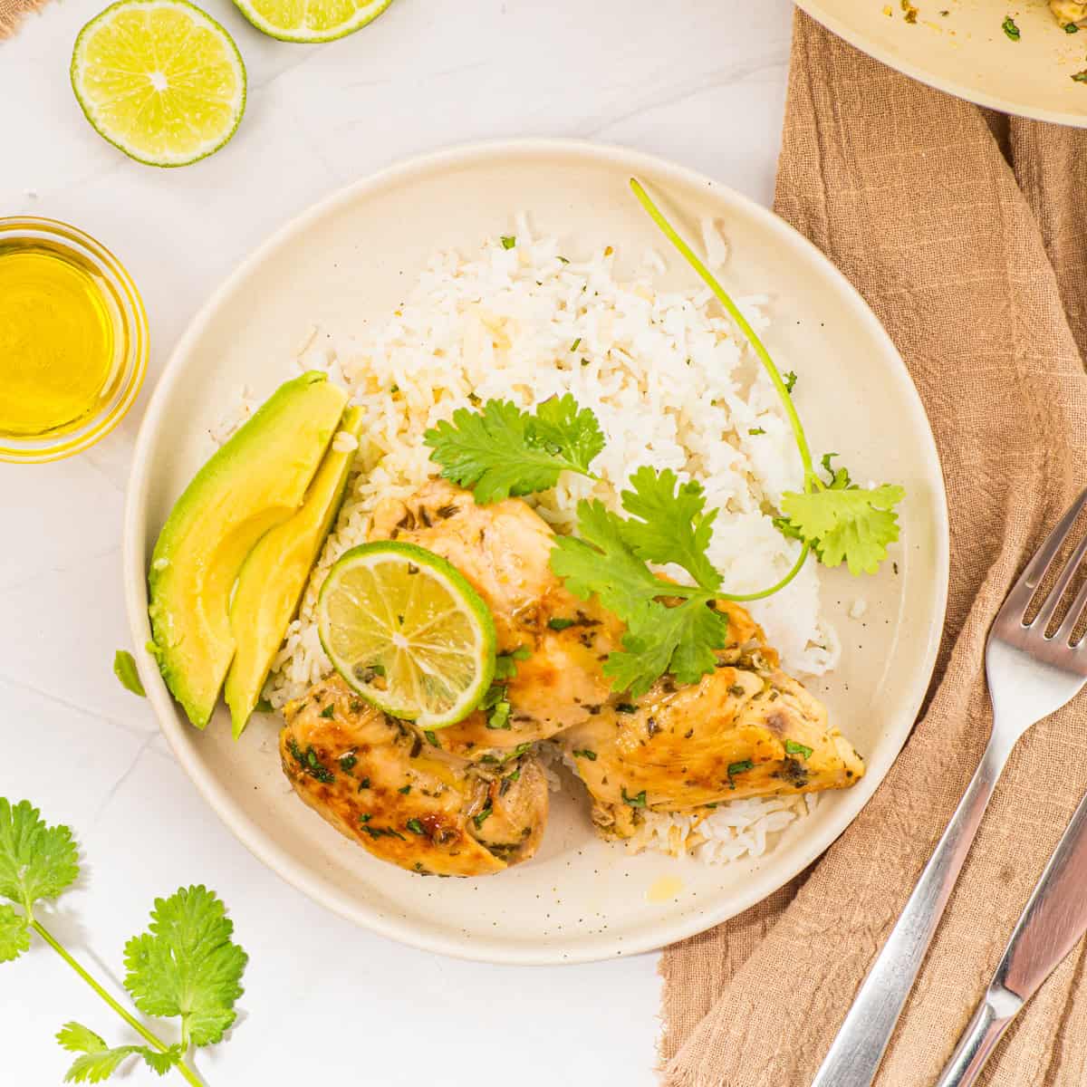 Plate with rice, grilled chicken topped with a lime slice and cilantro, avocado slices, a fork, a knife, and a brown napkin nearby.