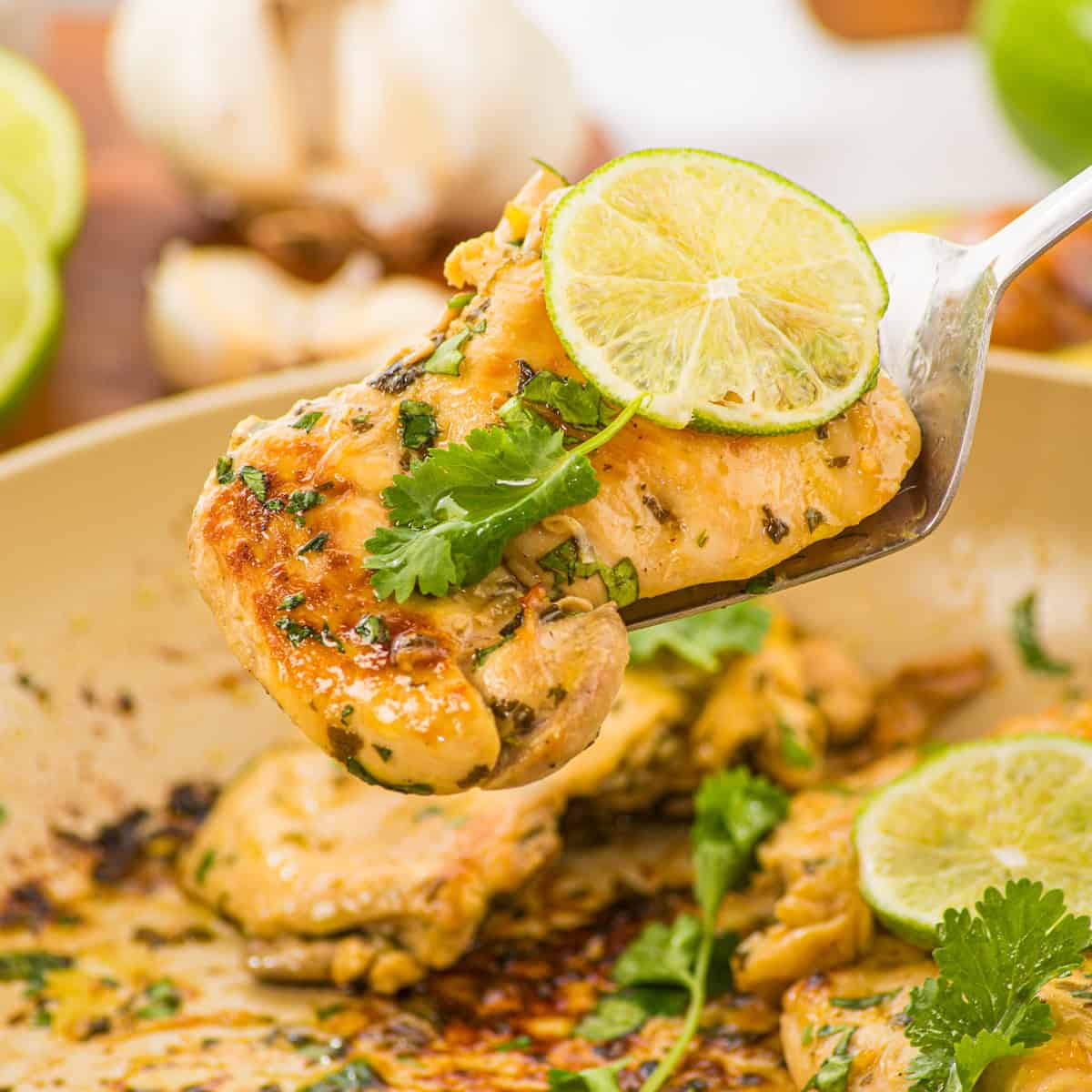 A fork holds a piece of cooked chicken garnished with cilantro and a slice of lime, with more chicken, lime, and garlic visible in the background.