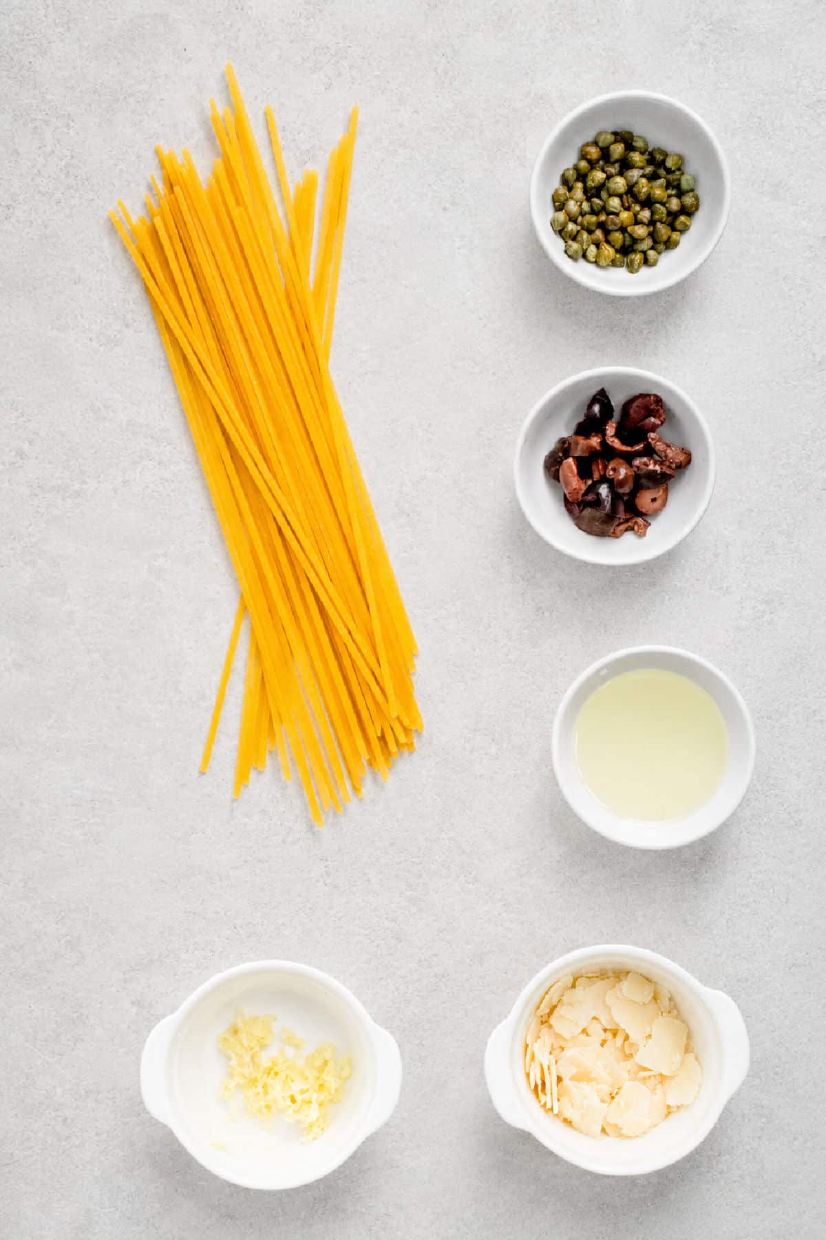 Uncooked pasta, capers, chopped olives, lemon juice, minced garlic, and shaved parmesan are arranged in separate white bowls on a light surface.