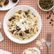 A plate of linguine pasta with olives, capers, and shaved parmesan on a red checkered tablecloth, surrounded by bread, extra parmesan, and a fork.