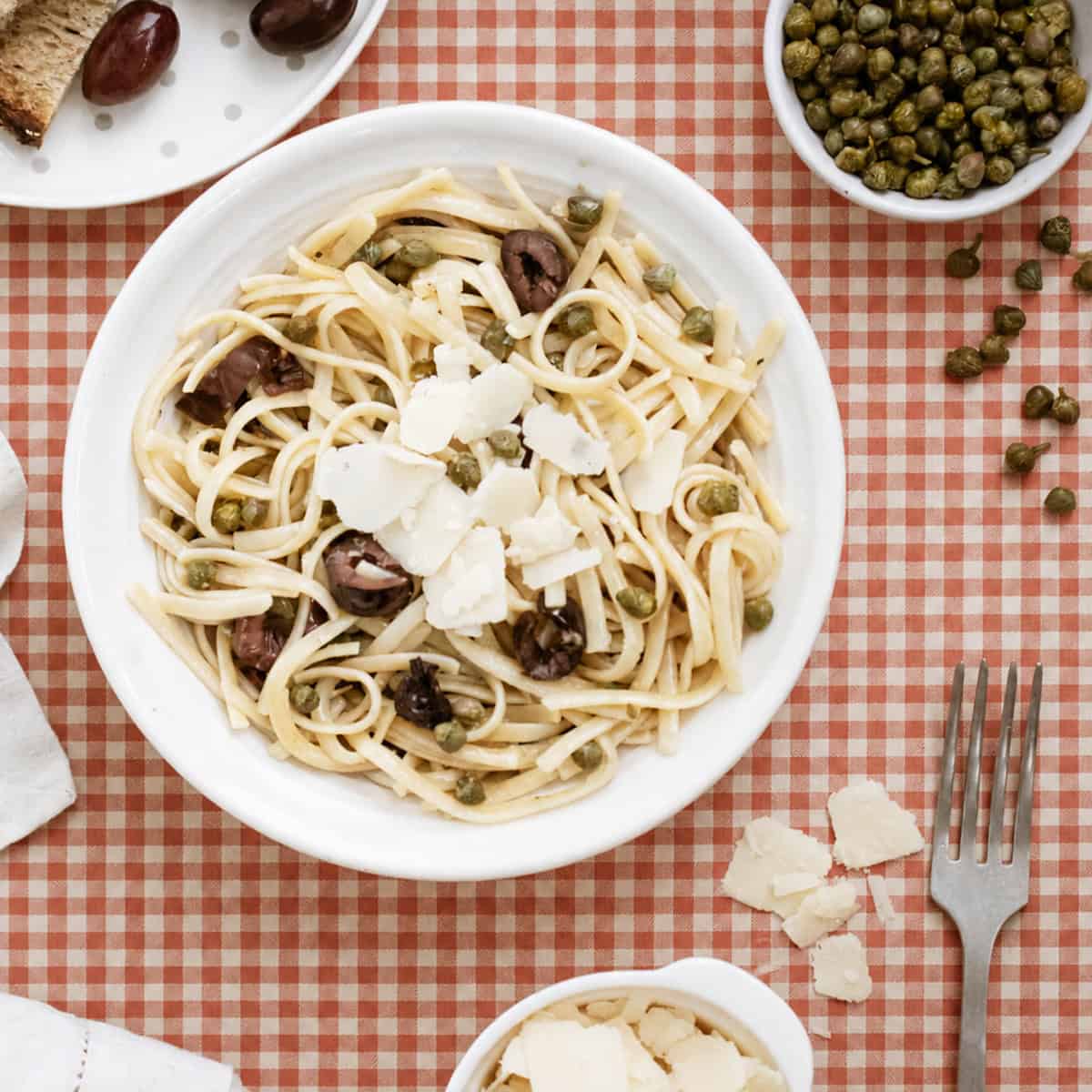A plate of linguine pasta with olives, capers, and shaved parmesan on a red checkered tablecloth, surrounded by bread, extra parmesan, and a fork.