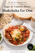 A plate of shakshuka with two eggs in tomato sauce, garnished with herbs, sits on a table next to bread slices, a fork, and a napkin.