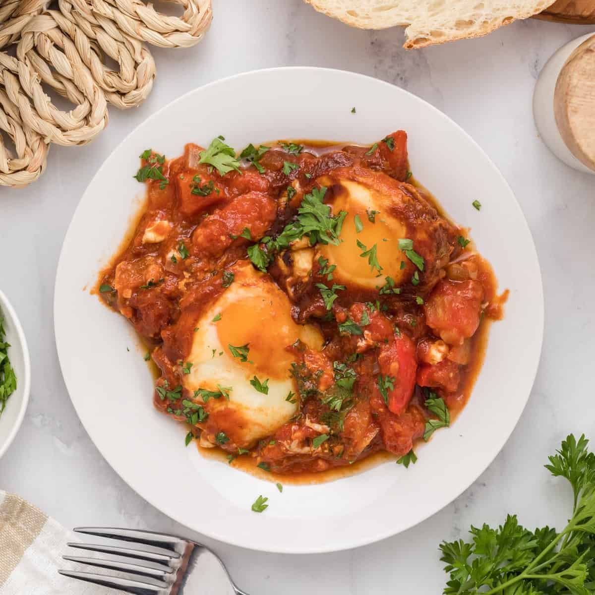 A white plate with shakshuka—poached eggs in a tomato sauce—garnished with chopped parsley, surrounded by bread and herbs on a white table.