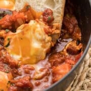 Eggs poached in tomato sauce with chopped vegetables and herbs, served in a black pan with a slice of bread dipped into the sauce.