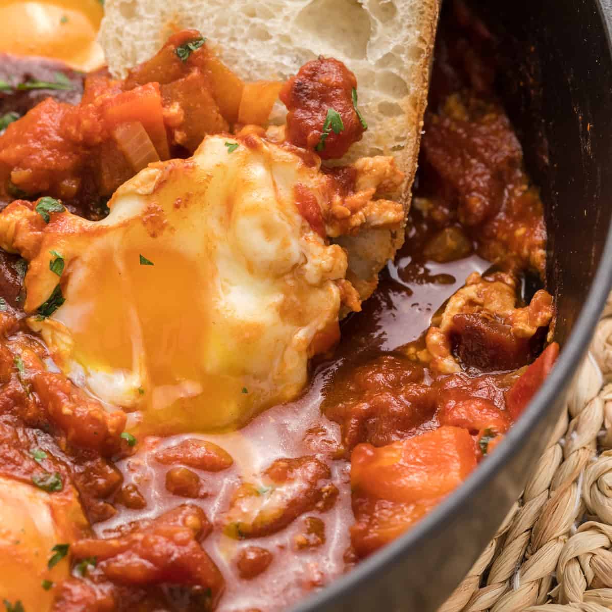 Eggs poached in tomato sauce with chopped vegetables and herbs, served in a black pan with a slice of bread dipped into the sauce.