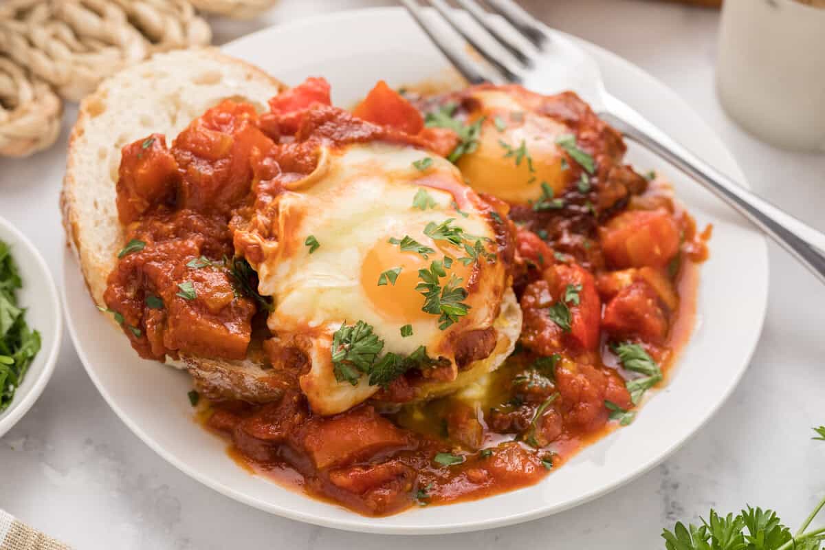 A plate of shakshuka with poached eggs in tomato sauce, garnished with parsley and served with a slice of bread and a fork.