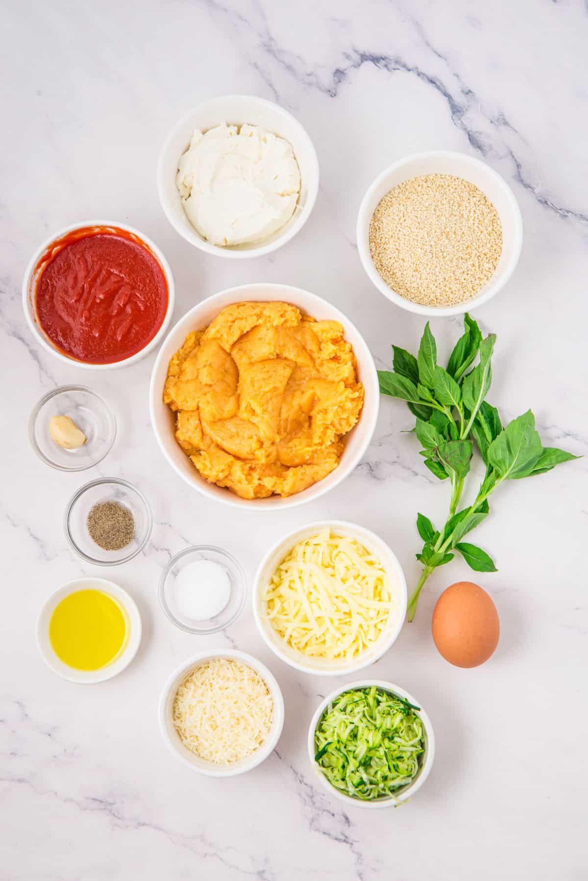 Various ingredients in bowls on a white surface, including marinara sauce, cheese, egg, basil, oil, garlic, spices, zucchini, ricotta, breadcrumbs, and ground chicken.