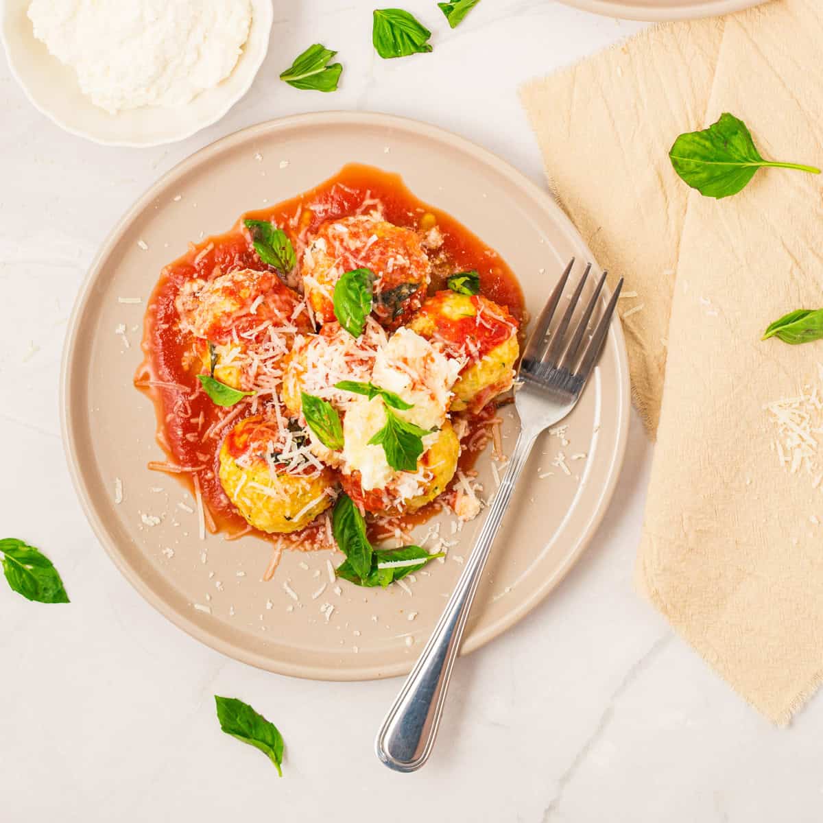 A beige plate with ricotta-stuffed pasta in tomato sauce, topped with grated cheese and fresh basil, sits beside a fork, napkin, and scattered basil leaves.