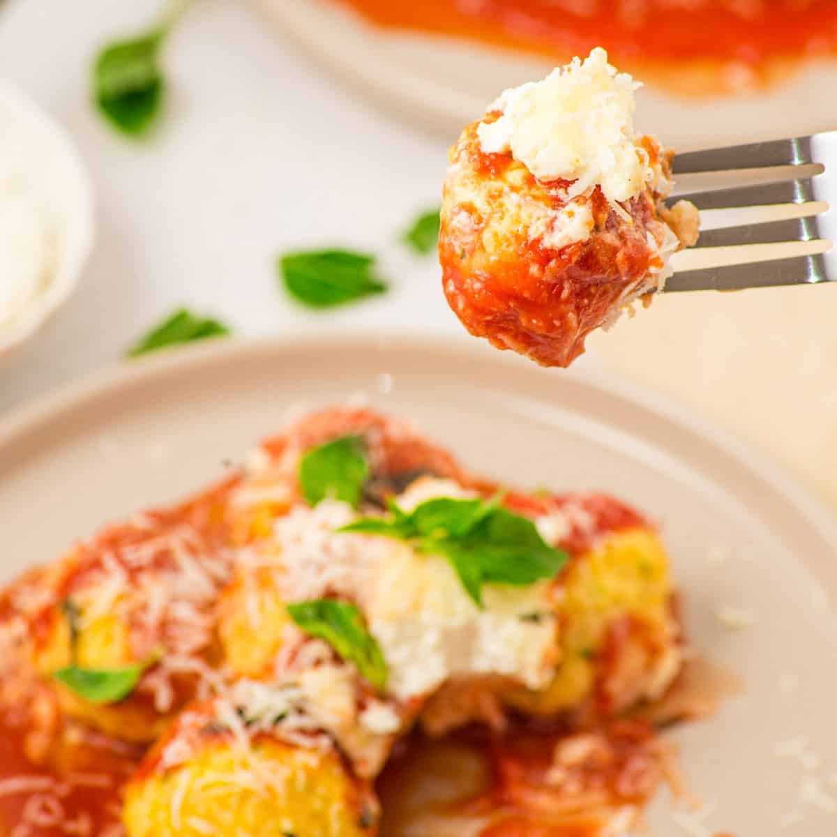 A fork holds a piece of ricotta and tomato sauce-topped baked manicotti above a plate, garnished with fresh basil and grated cheese.