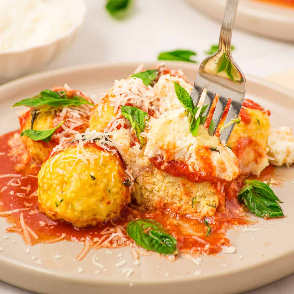 Four cheese-stuffed meatballs topped with tomato sauce, grated cheese, and fresh basil on a beige plate, with a fork cutting into one meatball.