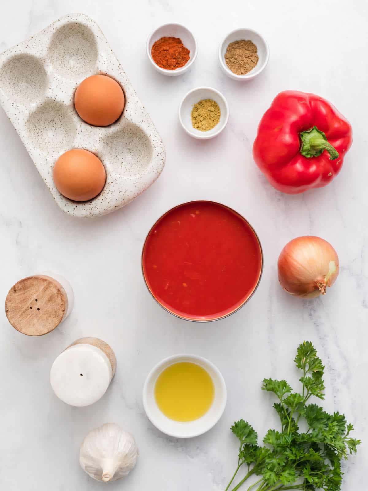 Eggs, spices, a red bell pepper, an onion, canned tomatoes, salt, pepper, garlic, olive oil, and parsley are arranged on a white surface—everything you need to make a delicious shakshuka for one.